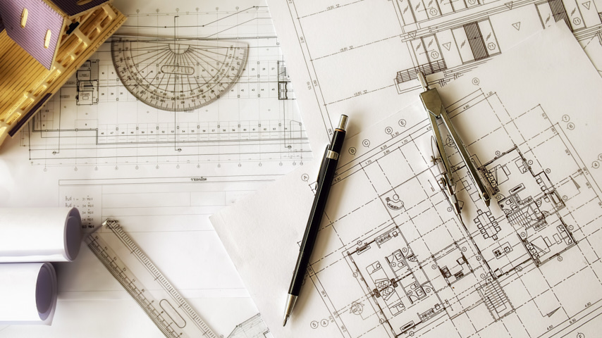 Sobre una mesa de trabajo planos abiertos, rollos de planos, compás, portaángulos y reglas